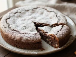Torta caprese al cioccolato e mandorle con spolverata di zucchero a velo e una fetta tagliata su un piatto di ceramica.