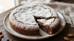 Torta caprese al cioccolato e mandorle con spolverata di zucchero a velo e una fetta tagliata su un piatto di ceramica.