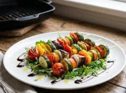Spiedini vegetariani grigliati serviti su un letto di rucola fresca in un piatto bianco, conditi con glassa di aceto balsamico e olio d'oliva aromatizzato.