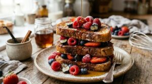 French Toast gourmet con frutti di bosco e sciroppo d'acero. Torre di French Toast dorati serviti su un piatto rustico, farciti con lamponi, mirtilli e fragole fresche, con sciroppo d'acero colato sopra.