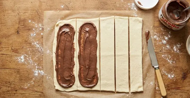 Spalmate la Nutella su due strisce di pasta sfoglia e ricopritele con le altre due strisce. Dividete ogni striscia a metà e poi dividete ogni metà verticalmente. Alla fine dovrete ottenere otto striscioline larghe circa 3 -4 centimetri.