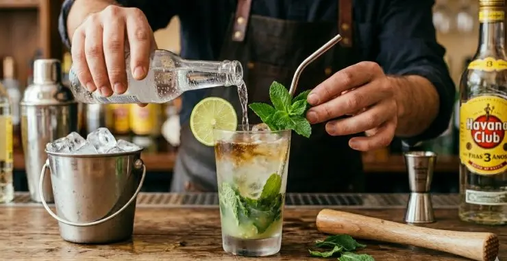 Riempite il bicchiere con l’acqua gassata, lasciando un centimetro di spazio dal bordo e versate in mezzo il resto del rum. Tagliate una fetta di lime e decorate il bicchiere. Aggiungete infine un rametto di menta e una cannuccia. Potete terminare la preparazione del mojito con 2 gocce di angostura (facoltative).