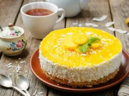 Ricetta Cheesecake al Mango Estiva Cheesecake al mango fresca senza cottura decorata con scaglie di cocco e menta su un tavolo in legno