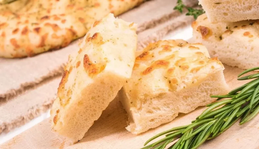 Fette di focaccia alta e soffice appena sfornata, disposte su un tagliere di legno accanto a un rametto di rosmarino fresco.
