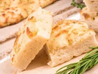Fette di focaccia alta e soffice appena sfornata, disposte su un tagliere di legno accanto a un rametto di rosmarino fresco.
