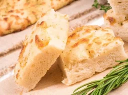Focaccia soffice fatta in casa con rosmarino Fette di focaccia alta e soffice appena sfornata, disposte su un tagliere di legno accanto a un rametto di rosmarino fresco.