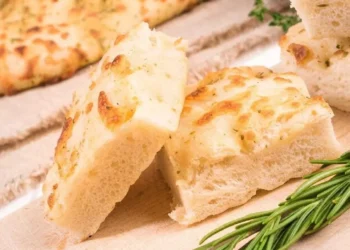 Fette di focaccia alta e soffice appena sfornata, disposte su un tagliere di legno accanto a un rametto di rosmarino fresco.