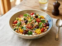 Ciotola centrale di insalata di ceci estiva con pomodorini, cetrioli, olive e basilico su tovaglia di lino.