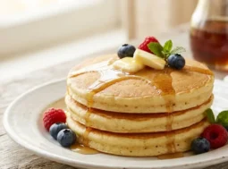 Pancake sofficissimi con 3 ingredienti Una pila di tre pancake soffici e alti guarniti con burro, sciroppo d'acero, mirtilli e lamponi freschi su un piatto bianco