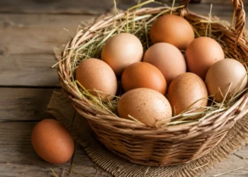Uova fresche in una cesta di vimini su tavolo rustico Cesta di vimini colma di uova fresche adagiate sulla paglia e appoggiata su un tavolo di legno rustico illuminato da luce naturale.