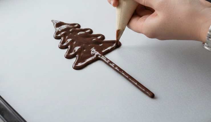 Andiamo a disegnare, premendo con delicatezza, un alberello dal basso verso l’alto sulla carta da forno: facciamo delle strisce da sinistra a destra, stringendole man mano che saliamo, senza mai interromperci. Una volta completato il disegno adagiamo uno stecchino di mikado da metà alberello a scendere verso la base che fungerà da tronco. Riprendiamo ora la sac a poche o la siringa e ricalchiamo l’intero albero, coprendo così anche la parte del mikado che si trova sul cioccolato e sigillando;