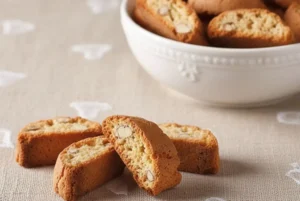 Cantucci toscani alle mandorle disposti su tovaglia rustica beige con cuori ricamati e ciotola bianca