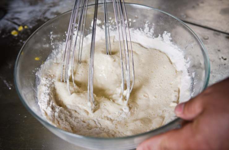 Accorpiamo la buccia di limone grattugiata e lo yogurt bianco. Lavoriamo ancora e aggiungiamo l’olio di semi. Mescoliamo ancora con le fruste e poi riversiamo nella crema anche il miele. Sbattiamo ancora e passiamo a setacciare le polveri: fatina e lievito;