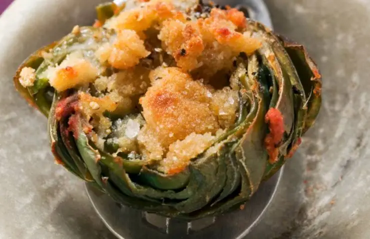 Oleiamo il fondo di una teglia da forno e distribuiamo i carciofi. Ricopriamo con il composto di parmigiano, pangrattato e aromi spolverando tutta la superficie. Inforniamo e cuociamo a 180 gradi per 20/30 minuti, fino a quando la gratinatura sarà ben dorata. Possiamo servire subito o lasciar intiepidire.