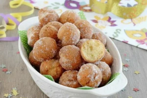 ricetta-castagnole-carnevale-originali Vassoio di castagnole fritte di Carnevale con zucchero, mascherina e stelle filanti su tavolo in legno.