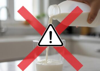 Una mano versa latte da una bottiglia bianca in un bicchiere di vetro, con un grande segnale di pericolo giallo sovrapposto per indicare un richiamo alimentare