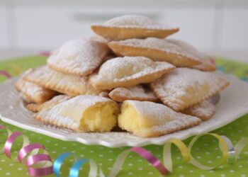 Un vassoio di porcellana bianca colmo di bugie di Carnevale ripiene di crema pasticcera, con due bugie aperte in primo piano che mostrano il ripieno vellutato, su una tovaglia verde a pois bianchi con stelle filanti.