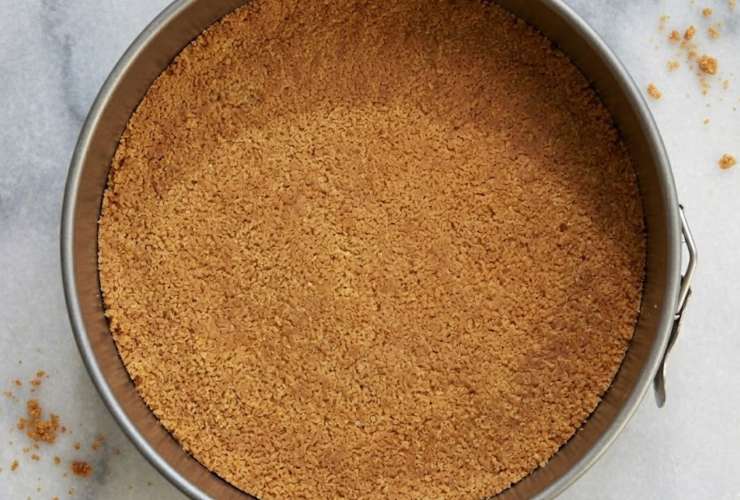 Uniamo i due ingredienti in una ciotola, amalgamiamo e riversiamo il composto ottenuto sul fondo di una tortiera con cerniera, lavorando con le mani per modellare, poi mettiamo in frigo;