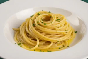 Primo piano di spaghetti aglio e olio cremosi con prezzemolo fresco in un piatto di porcellana bianca, su tovaglia verde a linee bianche con casseruola sullo sfondo.