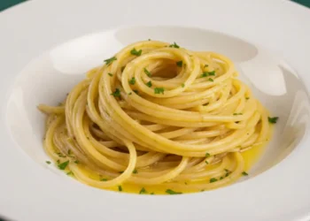 Primo piano di spaghetti aglio e olio cremosi con prezzemolo fresco in un piatto di porcellana bianca, su tovaglia verde a linee bianche con casseruola sullo sfondo.