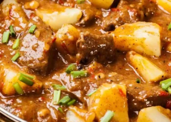 Ciotola bianca di spezzatino con patate tenerissimo fumante, servito su una tavola di legno bianco in una cucina luminosa stile shabby chic con una forchetta su un tovagliolo di lino