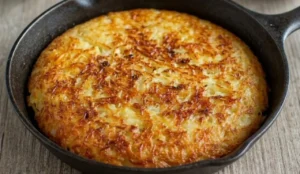 Frico friulano croccante alle patate e formaggio Montasio servito in una padella di ghisa nera su un tavolo di legno.