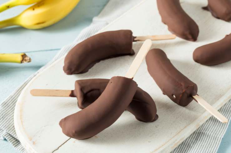 Immergiamo le banane nel cioccolato fuso reggendole dal lato dello spiedino ricoprendole per bene, quindi lasciamo rassodare il cioccolato il tempo necessario prima a temperatura e poi qualche minuto in frigo. Per ottenere una copertura spessa e golosa ripetiamo il passaggio un paio di volte. Non resta che decorare, se vogliamo e gustare.