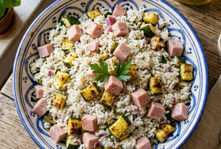 Scoliamo il riso, sgoccioliamo e riversiamo in una ciotola. Uniamo un filo d’olio d’oliva e mescoliamo lasciando raffreddare. Uniamo quindi le zucchine e il prosciutto a dadini, poi una spolverata di parmigiano grattugiato. Mescoliamo e aggiustiamo il sale, poi uniamo gli aromi che preferiamo: pronto da servire.