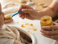 Mano di una mamma che imbocca un bambino con un omogeneizzato di patate e carote da un vasetto di vetro su un seggiolone.