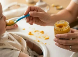 Mano di una mamma che imbocca un bambino con un omogeneizzato di patate e carote da un vasetto di vetro su un seggiolone.