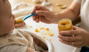 Mano di una mamma che imbocca un bambino con un omogeneizzato di patate e carote da un vasetto di vetro su un seggiolone.
