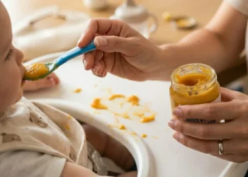 Mano di una mamma che imbocca un bambino con un omogeneizzato di patate e carote da un vasetto di vetro su un seggiolone.
