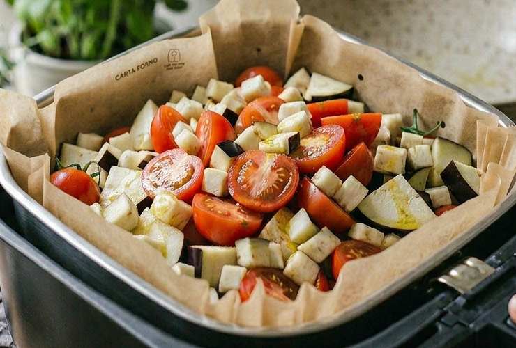 Foderiamo il cestello della friggitrice ad aria e riversiamo all’interno i pezzetti di melanzane e pomodori: non accavalliamoli troppo (procediamo in due cotture se la quantità è maggiore della capacità del cestello);