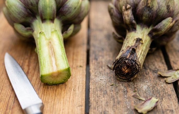 Primo piano ravvicinato di due gambi di carciofo a confronto sul banco del mercato. A sinistra, un taglio fresco, verde chiaro e umido; a destra, un taglio scuro, ossidato e floscio.