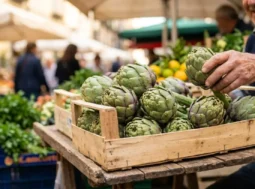 Una cassetta di legno colma di carciofi verdi sferici e turgidi su un banco del mercato all'aperto a Napoli, con sfondo sfocato e soleggiato.