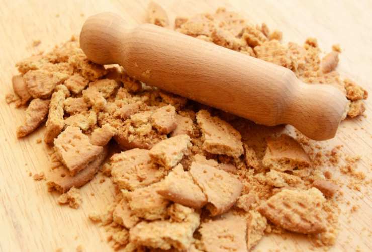 Iniziamo sbriciolando i biscotti alla meglio: possiamo usare un frullatore o un matterello, schiacciandoli dopo averli riversati in un sacchetto per alimenti. Intanto sciogliamo il burro a bagnomaria;