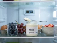 Vasetto di yogurt naturale aperto in un frigorifero organizzato a 3.8°C con cucchiaio e mirtilli freschi.