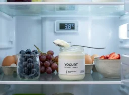 Vasetto di yogurt naturale aperto in un frigorifero organizzato a 3.8°C con cucchiaio e mirtilli freschi.