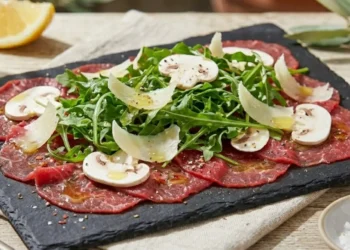 Fettine di carpaccio di manzo rosso vivo con rucola turgida, scaglie di grana e funghi champignon sodi su un piatto di ardesia.