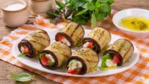 Involtini di melanzane grigliate lucidi di olio extravergine d'oliva con ripieno compatto di feta, pomodorini e basilico, disposti su un piatto bianco sopra uno strofinaccio a scacchi arancioni.