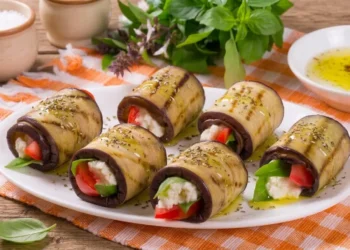 Involtini di melanzane grigliate lucidi di olio extravergine d'oliva con ripieno compatto di feta, pomodorini e basilico, disposti su un piatto bianco sopra uno strofinaccio a scacchi arancioni.