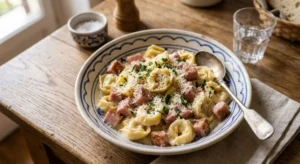 Un piatto di ceramica con tortellini panna e prosciutto cotto a cubetti, spolverati con formaggio grattugiato, pepe nero e prezzemolo fresco, servito su un tavolo di legno rustico.