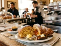 Un cornetto al bar tagliato a metà che mostra il ripieno di marmellata e la sfogliatura interna, appoggiato sul bancone di una caffetteria italiana con una tazzina di caffè espresso e macchina del caffè sullo sfondo sfocato.