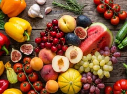 Composizione dall'alto di frutta e verdura estiva di stagione su un tavolo in legno scuro, con anguria, melone, pomodori e peperoni colorati.