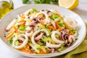 Insalata di calamari con verdure croccanti e limone Insalata di calamari teneri con sedano croccante, carote julienne, prezzemolo fresco e citronette al limone servita in una ciotola bianca.