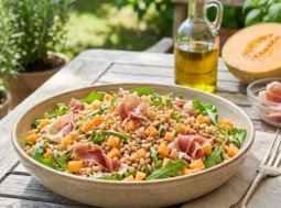Insalata di Farro Prosciutto e Melone Un'insalata di farro fredda servita in una ciotola con cubetti di melone, rucola fresca e straccetti di prosciutto crudo su un tavolo di legno all'aperto.