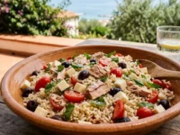 Insalata di riso mediterranea sgranata con filetti di tonno, pomodorini, olive nere e cubetti di primosale, servita in una ciotola di terracotta su un tavolo di legno all'aperto.