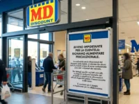 Ingresso di un supermercato MD con un grande cartello informativo in primo piano che segnala un avviso importante di richiamo alimentare precauzionale per la clientela.