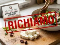 Insalata in preparazione in una ciotola con cubetti di formaggio feta sul tagliere di legno in primo piano, sormontata da un timbro grafico rosso con la scritta RICHIAMO.