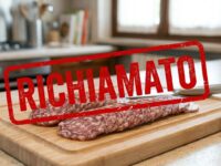 Primo piano di un salame tagliato a fette su un tagliere di legno in cucina, sormontato da una scritta rossa grafica a forma di timbro con il testo RICHIAMATO.